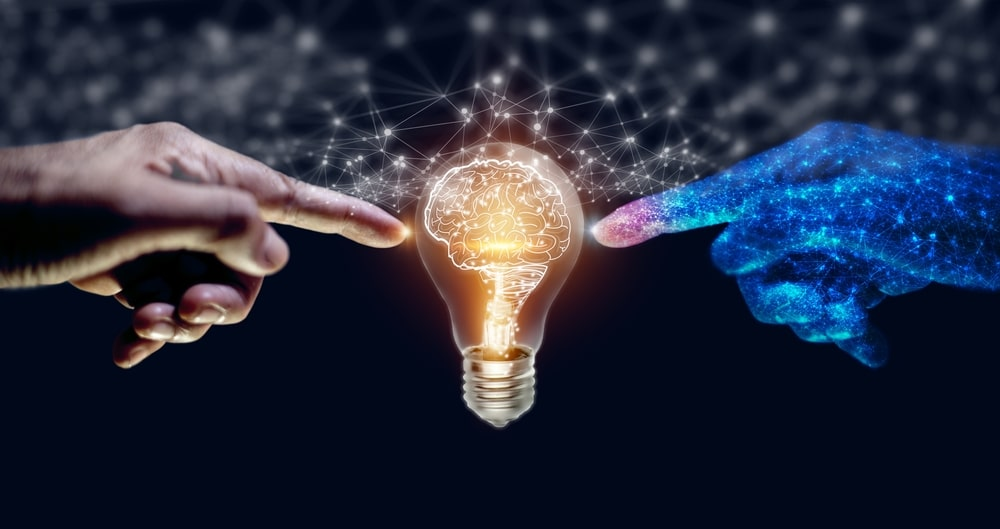 Uma mão humana tocando uma lâmpada que indica novas ideias junto a uma mão robótica, resumindo o encontro dos seres humanos e da inteligência artificial nessa busca incessante pelo novo.