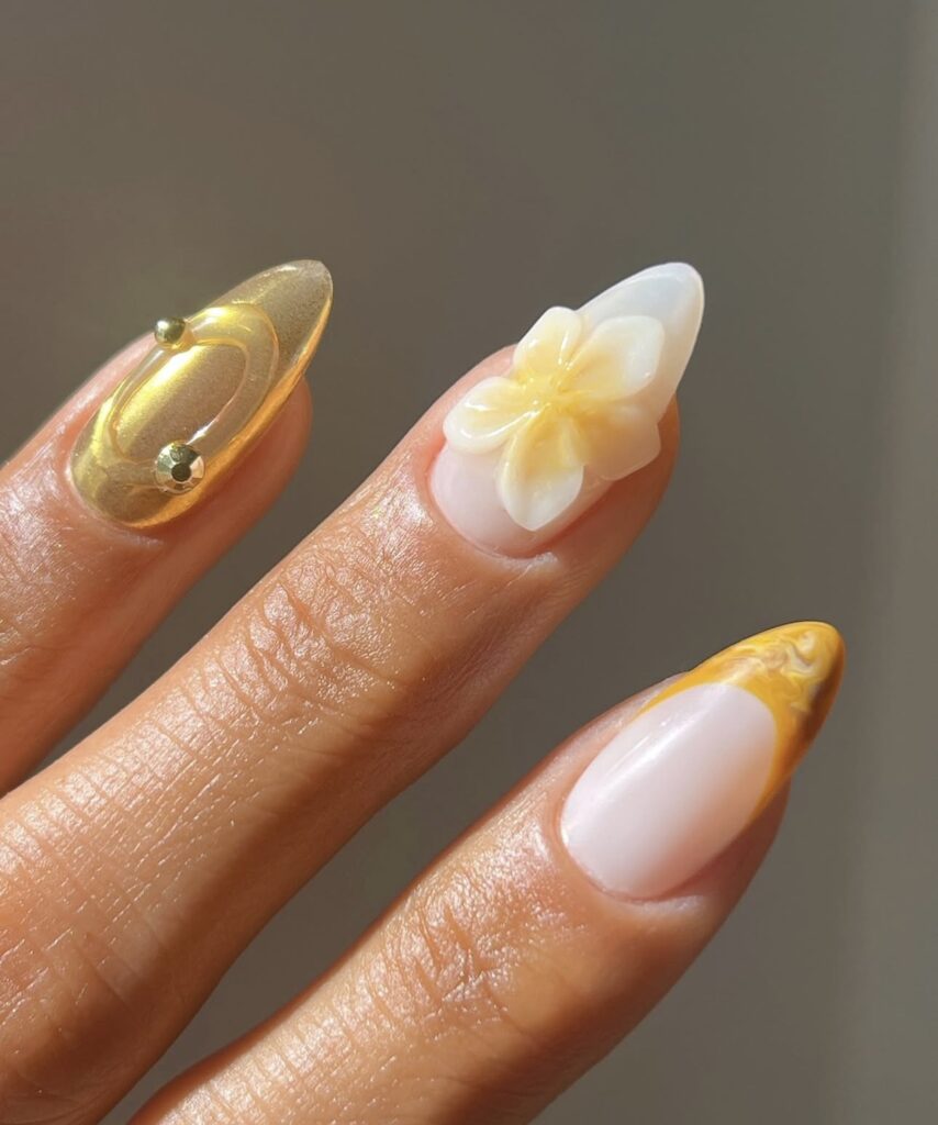 Exemplo de uma das tendências em alongamentos de unhas citadas neste post, arte 3d com unhas em formato stiletto com decoração sutil em tons de branco, dourado e amarelo, com elementos tridimensionais como aplicações de florais realistas
