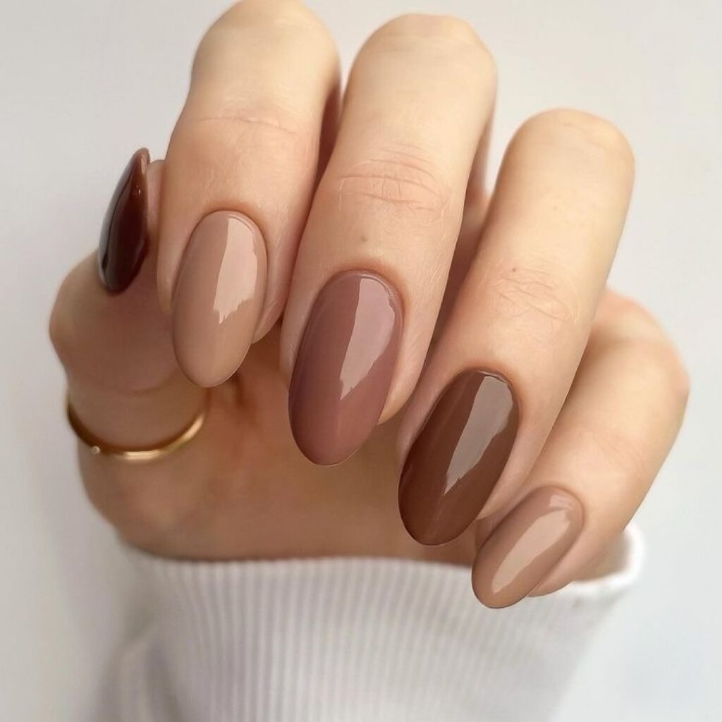 Exemplo de uma das tendências em alongamentos de unhas citadas neste post, unhas em formato almond sem decoração, porém há pintura nas unhas, com foco em tons de nude e terrosos conforme tendência mocha mousse. Além de anéis para decorar.