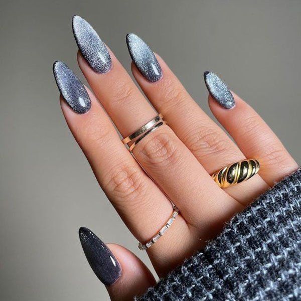 Exemplo de uma das tendências em alongamentos de unhas citadas neste post, conhecida como velvet nails. Mão com unhas em formato de stiletto, em uma paleta de tons metálicos e sombreados. Além de anéis para decorar.